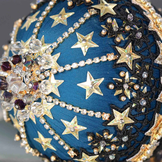 A close-up of dark purple beads and gold star-shaped sequins on a dark teal satin foam core ball featuring a completed handmade push pin ornament set against a dark gray background.