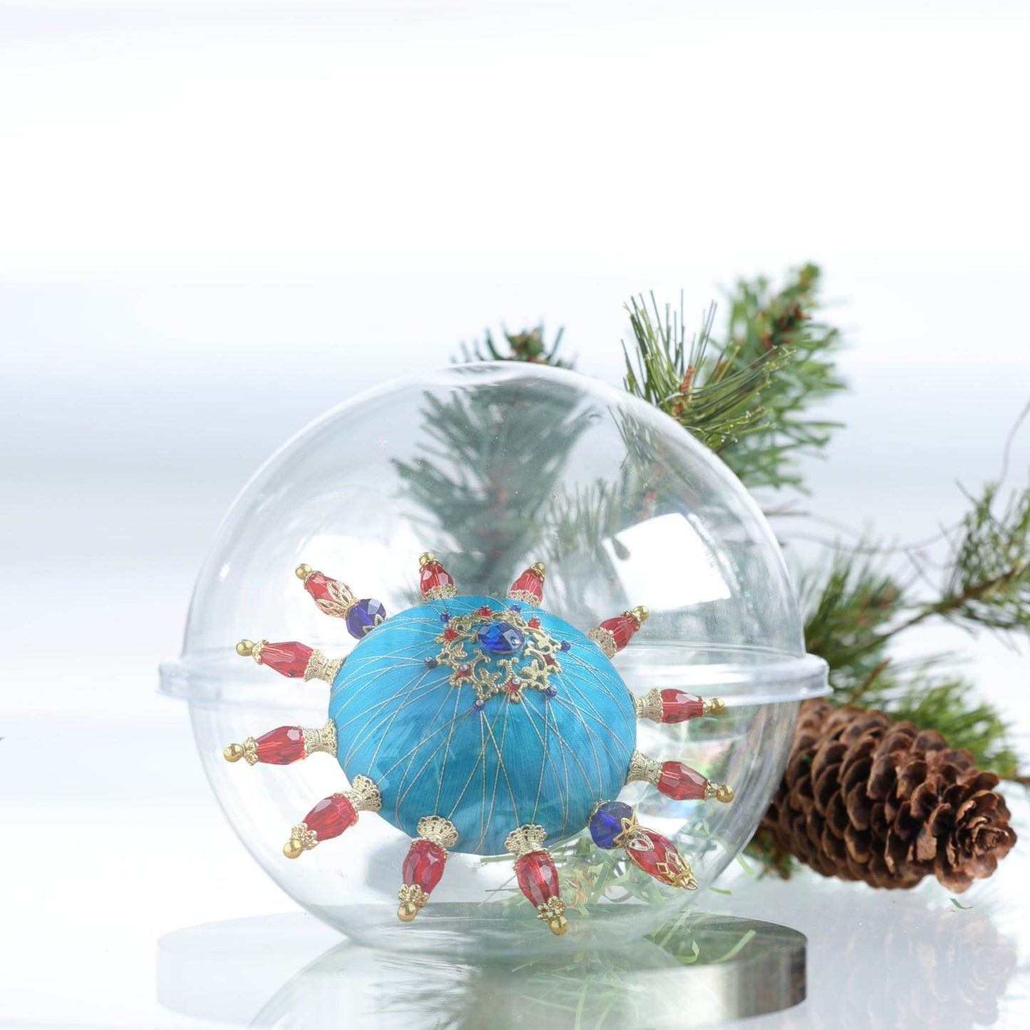 Christmas blue handmade ornament with red and gold accents inside a clear glass storage container, surrounded by pine branches and a pine cone by Whim5seasons.