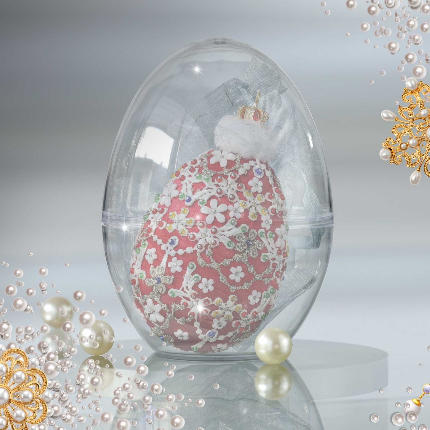 A floral Easter egg-shaped satin beaded ornament displayed inside a clear plastic egg-shaped container, with pearl-like props and ornate gold floral decorations around it on a light gray reflective background by Whim5seasons.