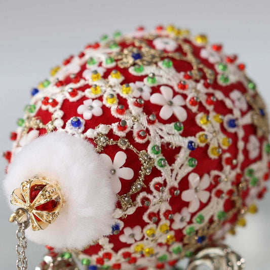 A close-up of an ornate red Christmas push pin ornament decorated with white floral embroidery, multicolored rhinestones, gold trim, and a fluffy white pom-pom with a gold crown by Whim5seasons.