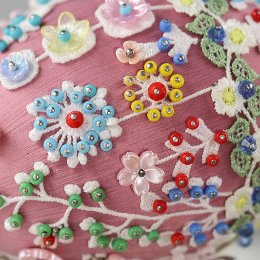 A close-up detail of a mauve Easter egg push pin ornament showing small beads pinned with dressmaker pins to the white lace by Whim5seasons.