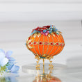 Load image into Gallery viewer, A side detail of a handmade orange Easter egg satin ornament with embellishments including red rhinestones, gold trim, and beads. The ornament is displayed on a gold stand.