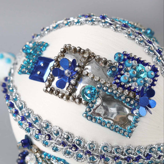 Close up of white handmade Christmas beaded push pin ornament in with sequined blue gift boxes and a snowman porcelain bead in the background.