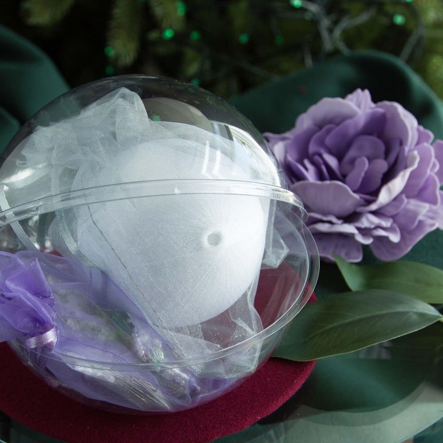 White satin ball with sheer taffeta bag, clear container, surrounded by purple flowers and green leaves. Close-up detail of Satin Handmade Ornament Trellis item with colorful flowers and a pearl on a white. (White).