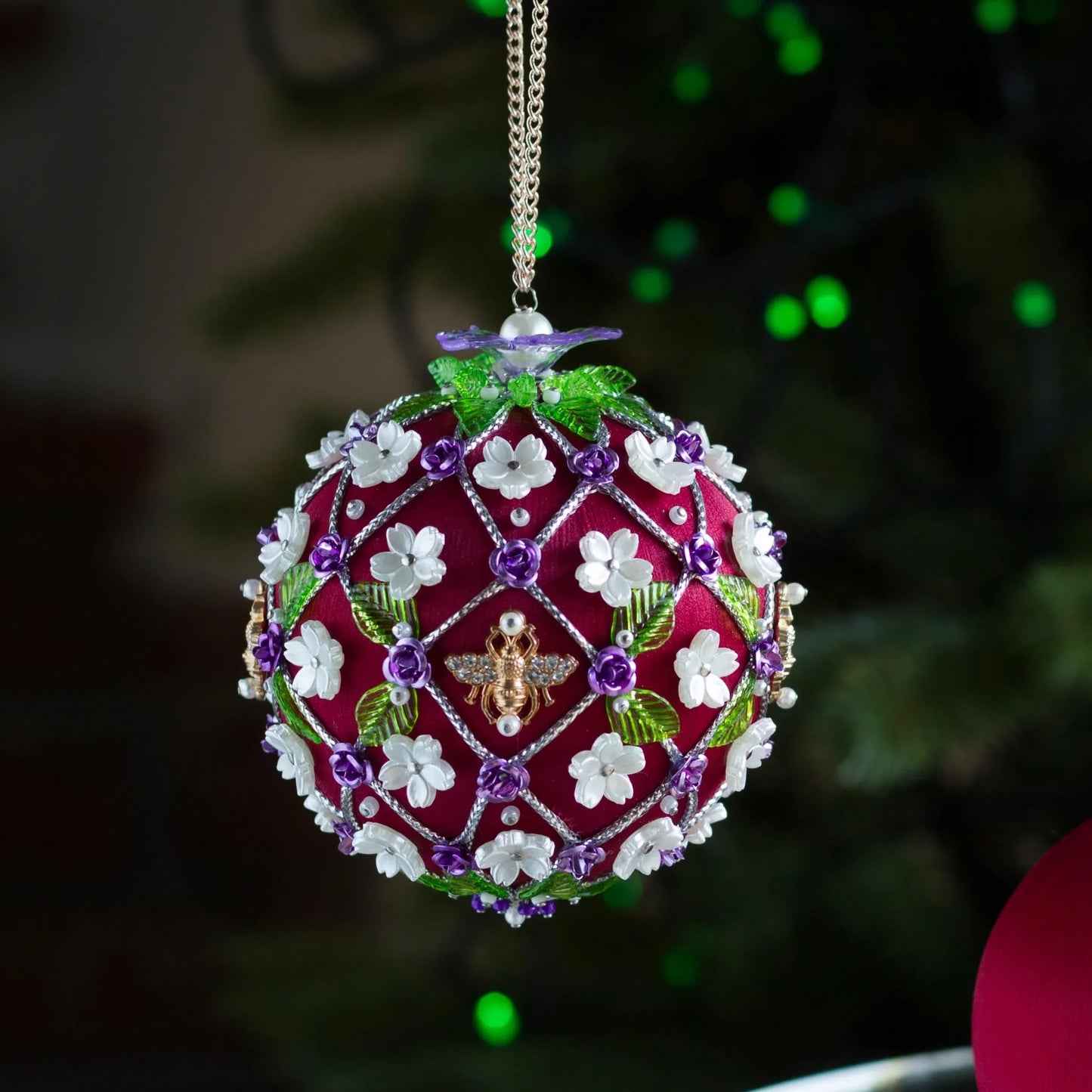 Decorative Handmade Christmas ornament with floral patterns and a bee design, hanging against a blurred green background. (Dark Red).