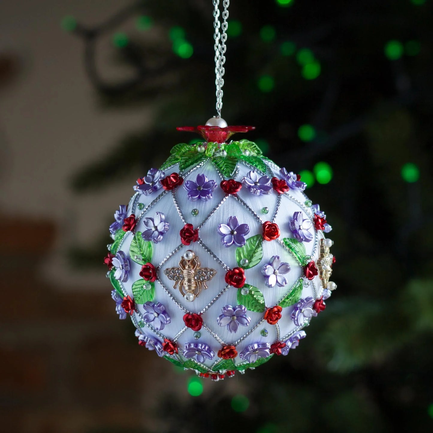Christmas handmade beaded ornament with floral patterns and a bee design, hanging against a blurred green background. (White).