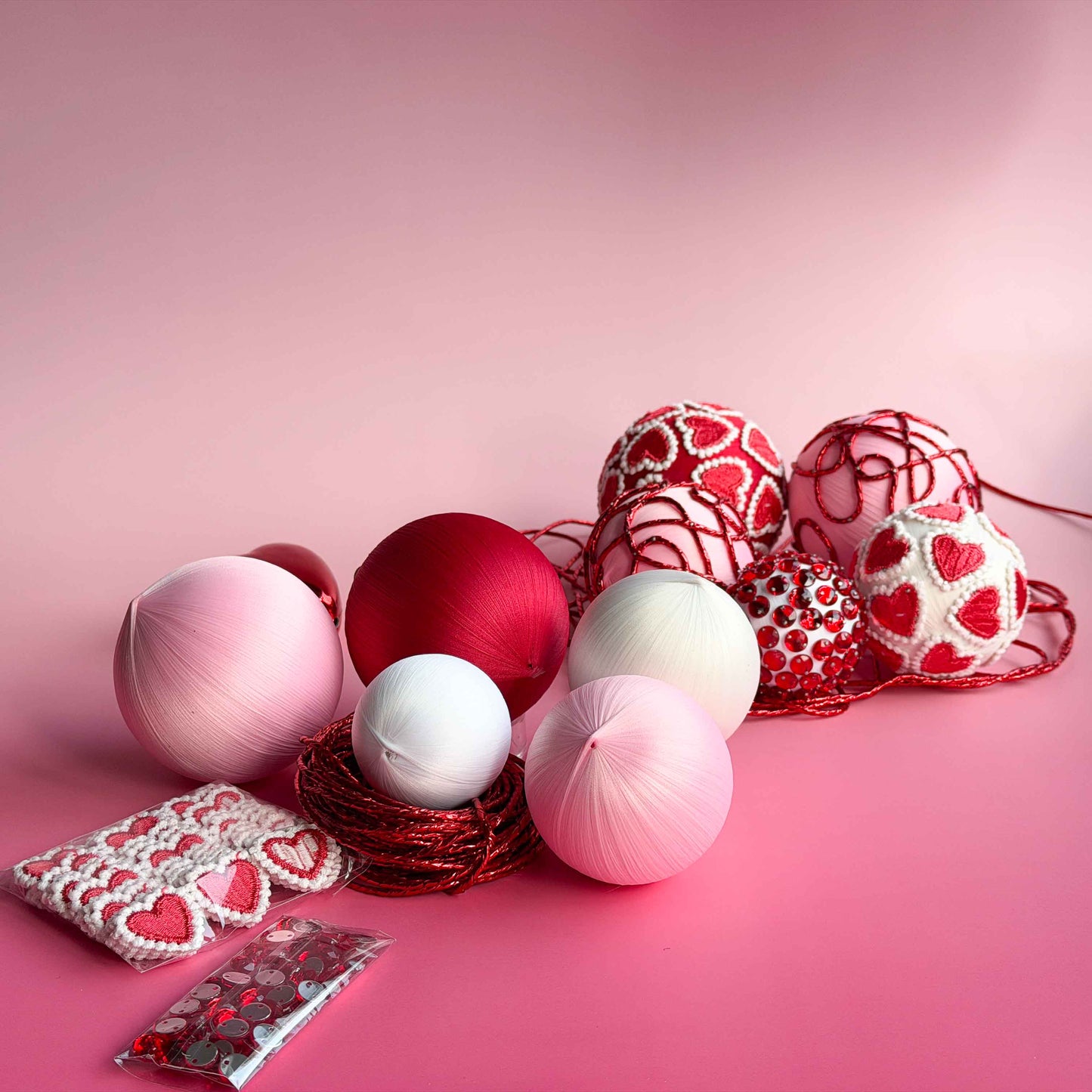 Materials for a Valentines Day DIY projects include heart-patterned trims, satin foam core balls, pins, and a small pouch of red rhinestones on a pink background by Whim5seasons.