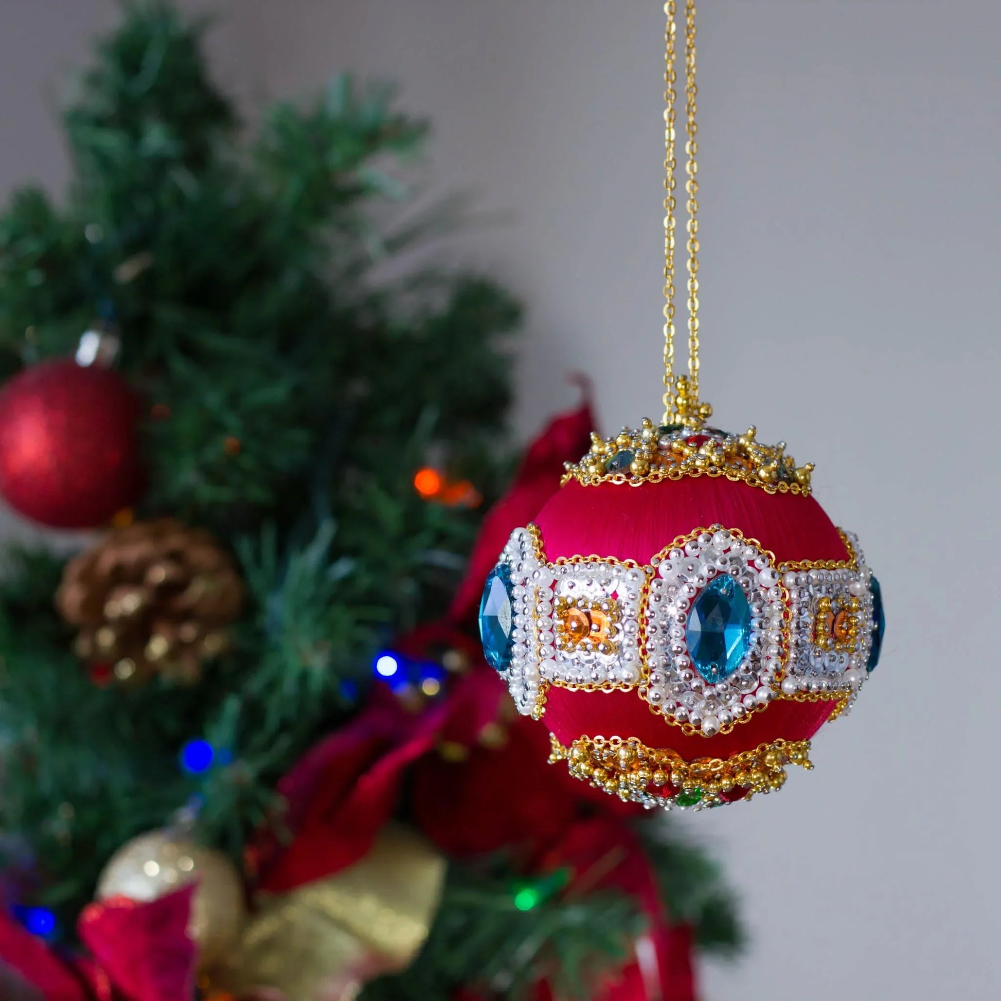 Gusornament Satin Ornament -Dark red beaded Christmas Ornament, embellished with blue glass rhinestones and hundreds of pearls and sequins, in front of a festive tree.  (Dark Red)