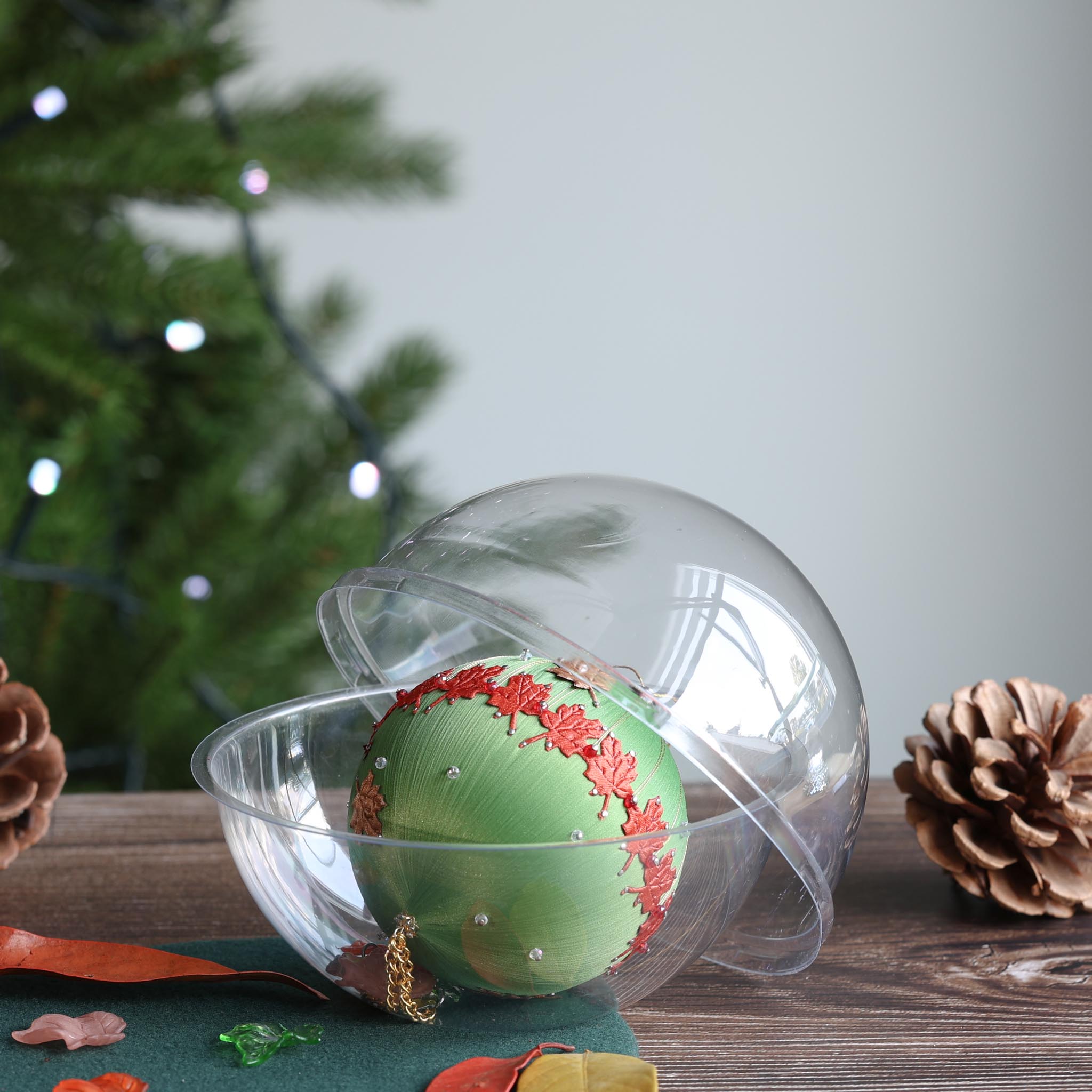Green satin ball with a kit in a clear plastic round container on a wooden surface with decorative elements and a blurred Christmas tree in the background. (Olive)