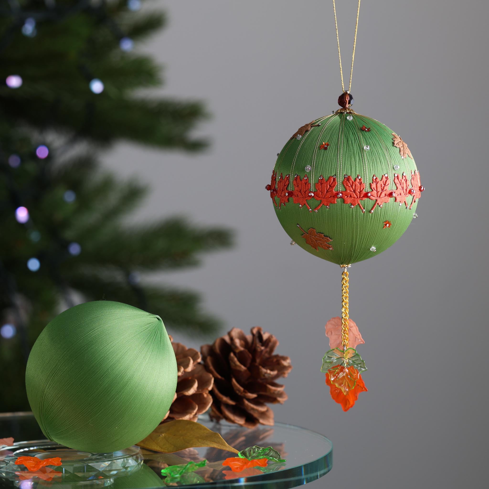 Green ornament kit with red maple leaf trim in front of a Christmas tree. (Olive)
