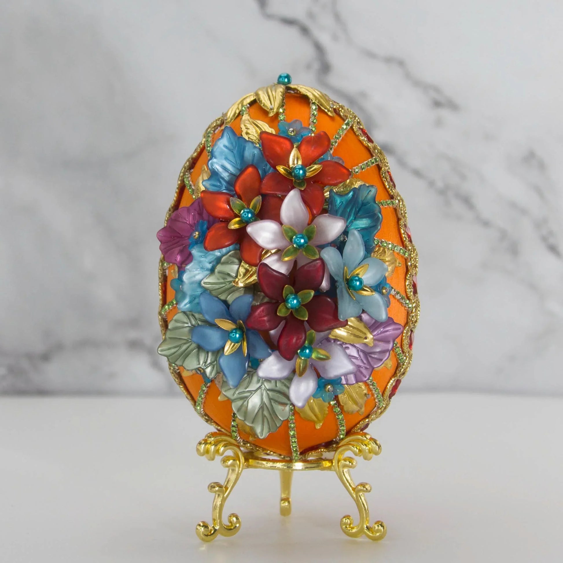 A handmade large Easter egg beaded ornament in gold and orange with embellishments including pins, rhinestones, and beads. The ornament is displayed on a gold stand.
