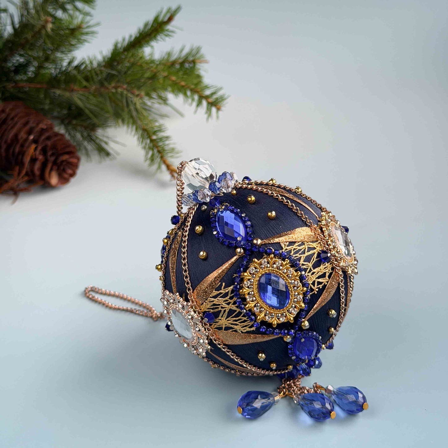 Decorative handmade dark blue push pin Christmas ornament with blue and gold details on a light blue background with an evergreen branch by Whim5seasons. (Navy)