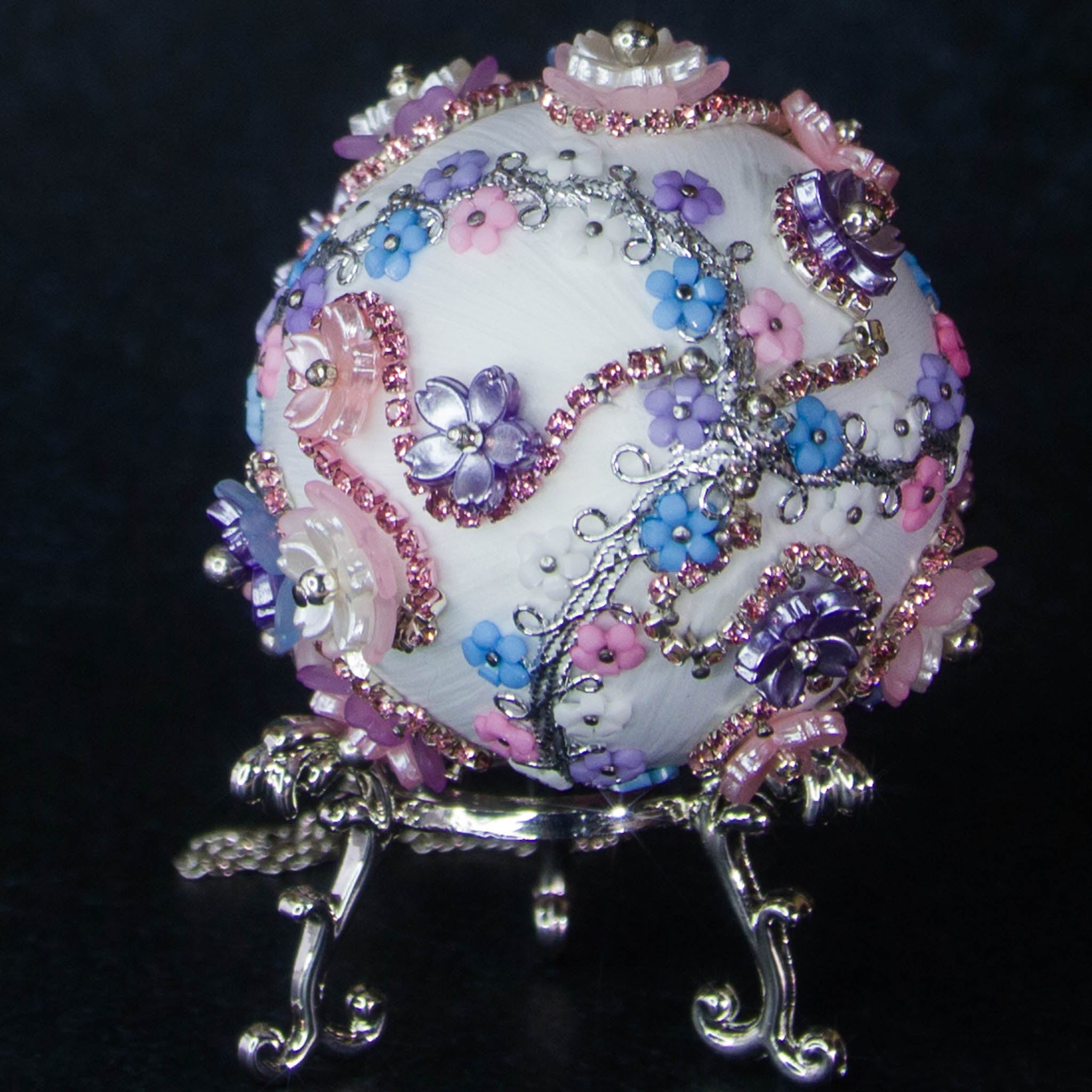 Princess Satin Egg Ornament -a close-up of an elegant Handmade Easter decoration, sitting on a display stand, adorned with sparkling beads, trims, rhinestones, and daisies, set against a dark background.