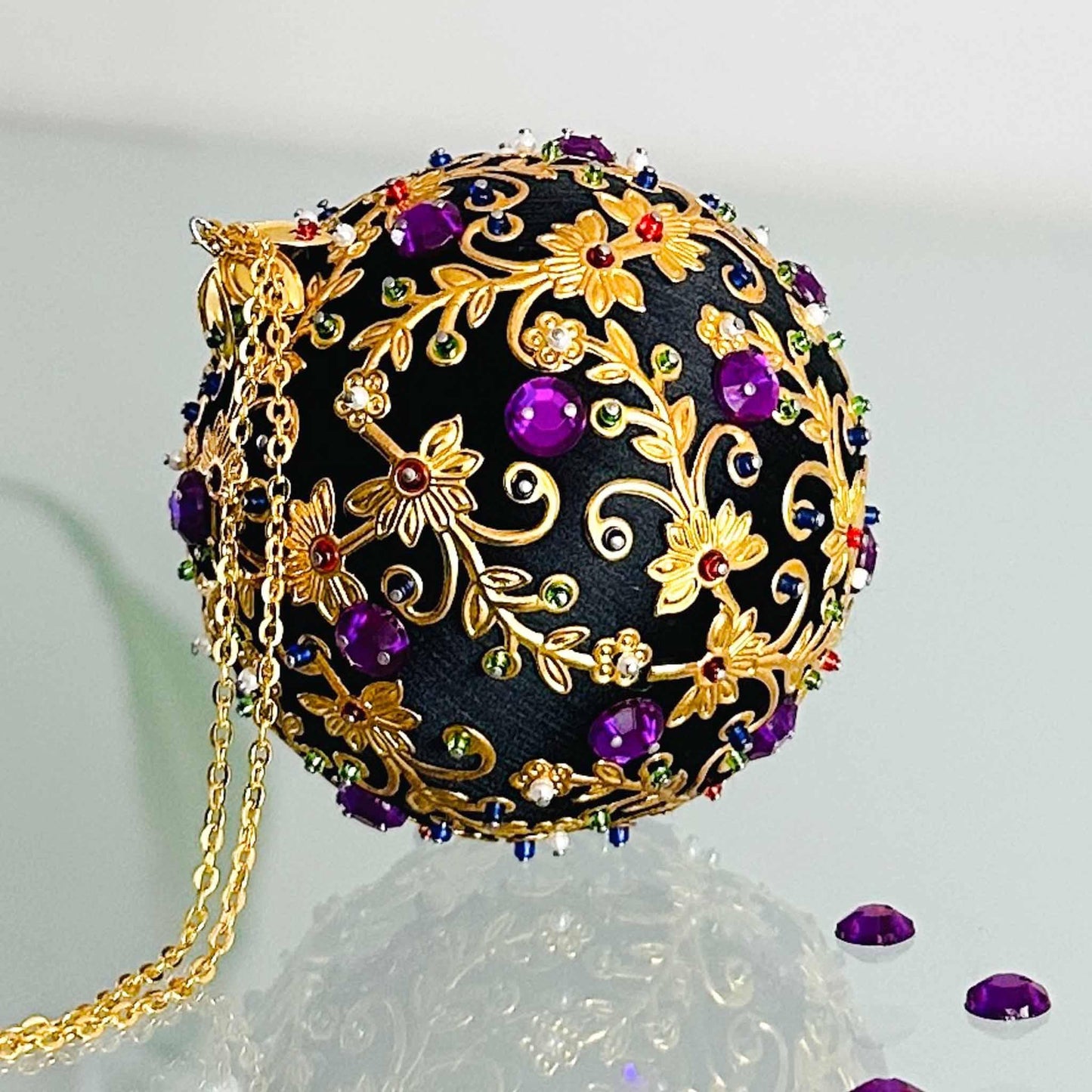 Quam Handmade Christmas Satin Ornament in Black with gold metal embellishments and purple rhinestones in front of evergreen branch. (Black)