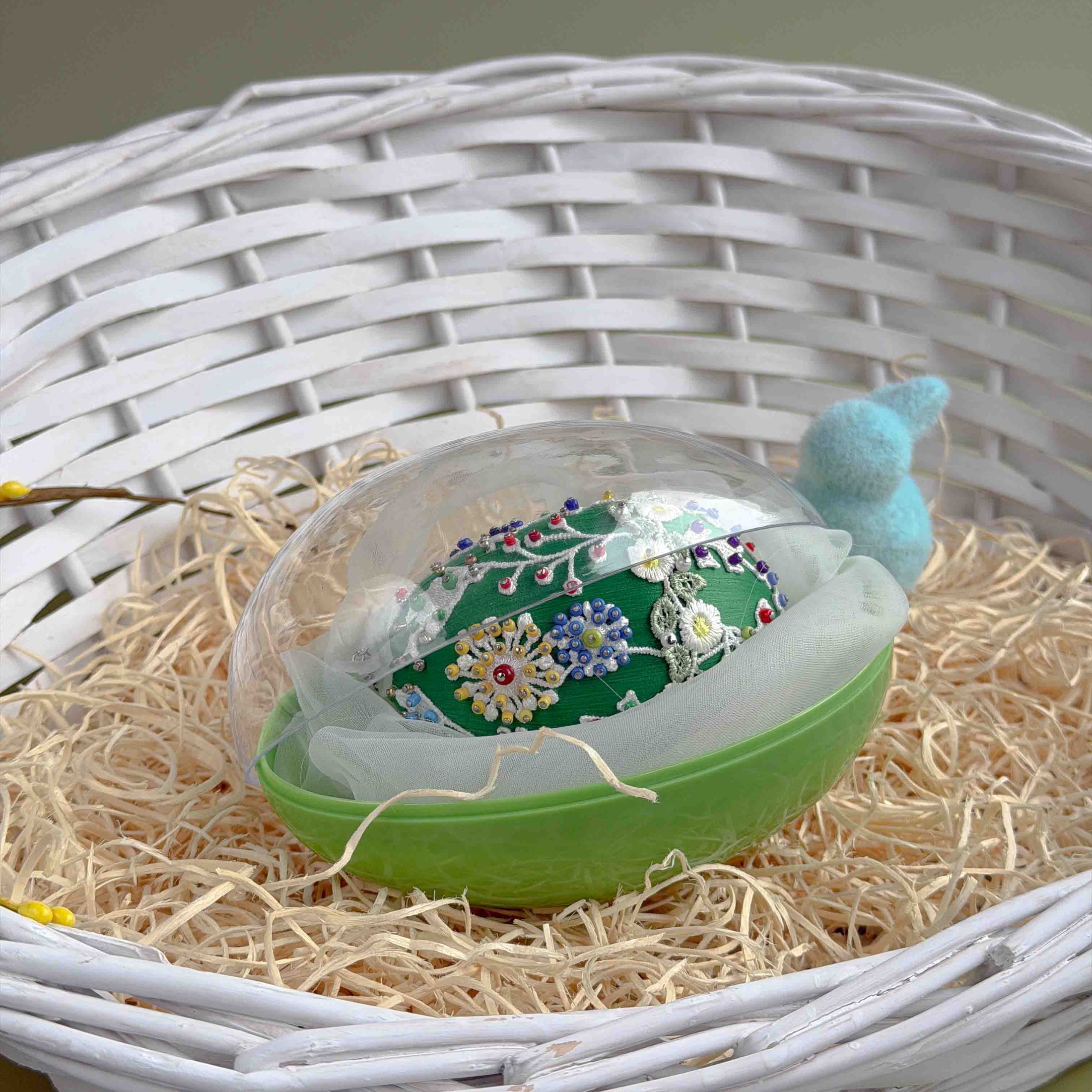 Decorative Easter egg with floral design in a green holder on straw inside a white wicker basket. (Green)