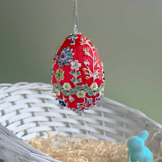 Decorative red egg with floral patterns hanging on a chain, placed in a woven basket with straw. (Dark Red)
