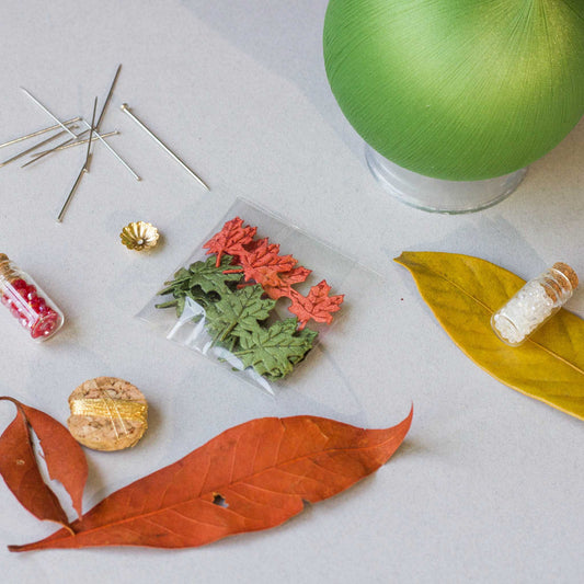 Push Pin Ornament includes these materials: assorted maple leaf charms, leaf ribbon, beads, and gold thread, on a light surface. (Olive)