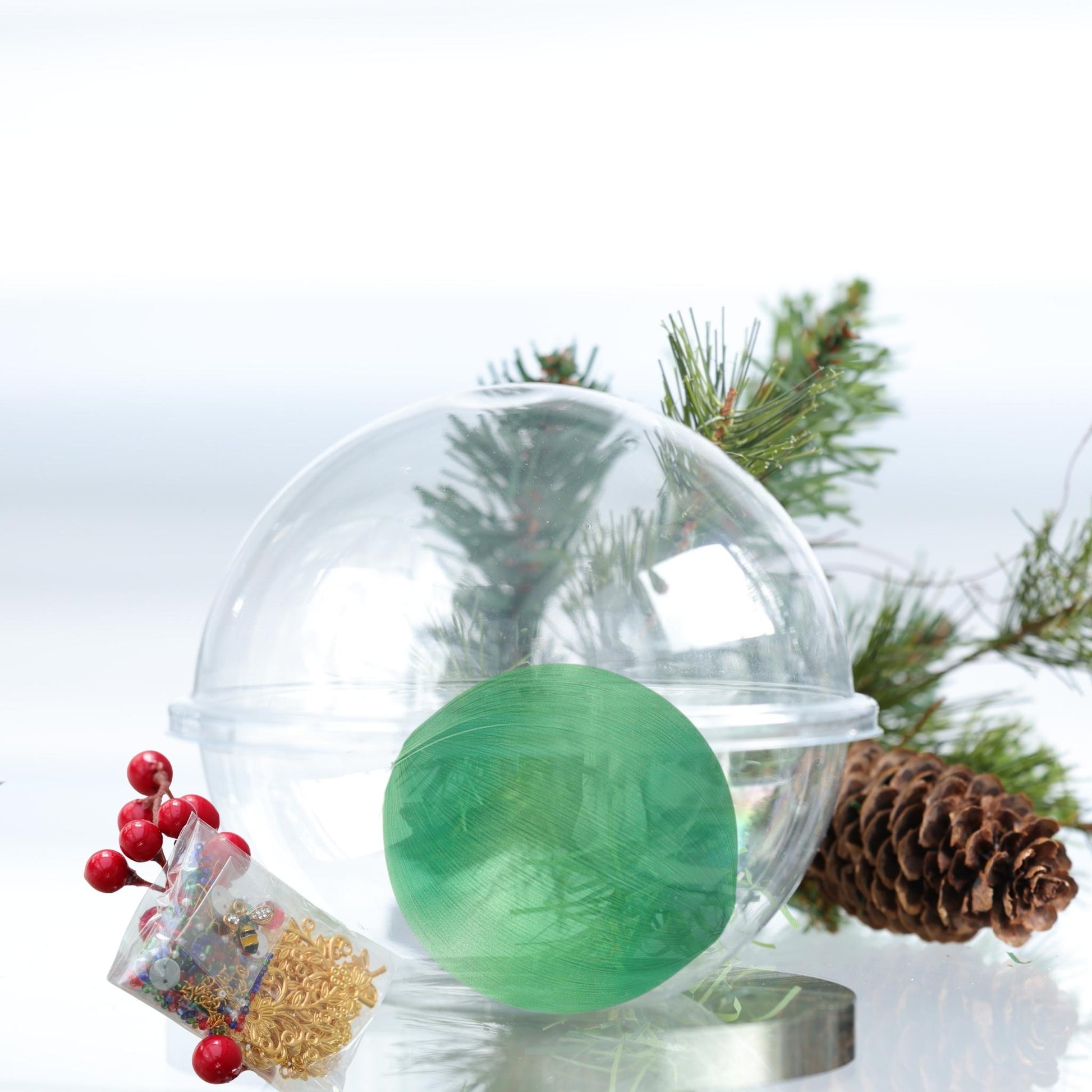 Green satin ball in a clear plastic storage container with Christmas decorations on a white background and a small package containing push pin kit ingridients by Whim5seasons. 