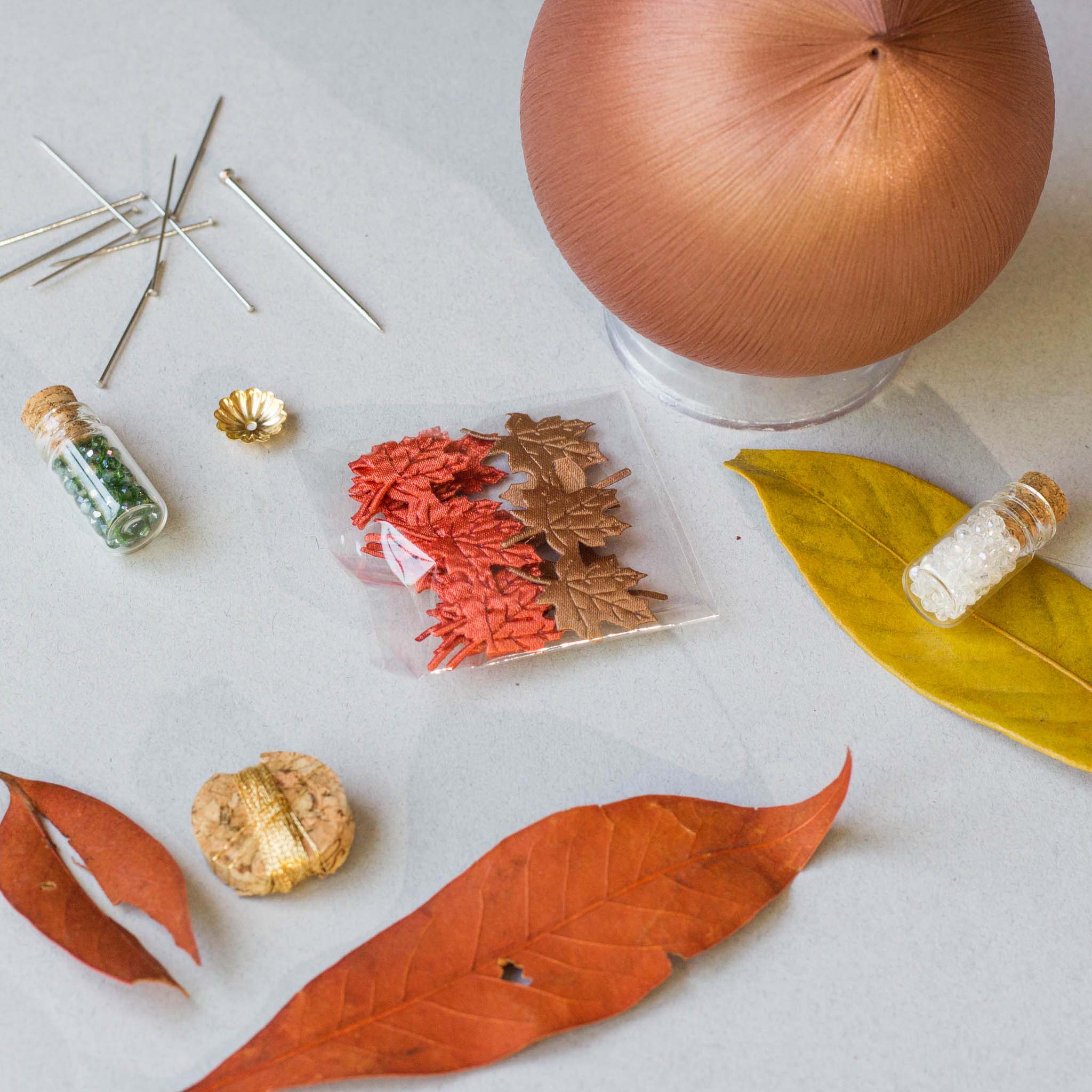 Push Pin Ornament includes these materials: assorted maple leaf charms, leaf ribbon, beads, and gold thread, on a light surface. (RosyBrown)
