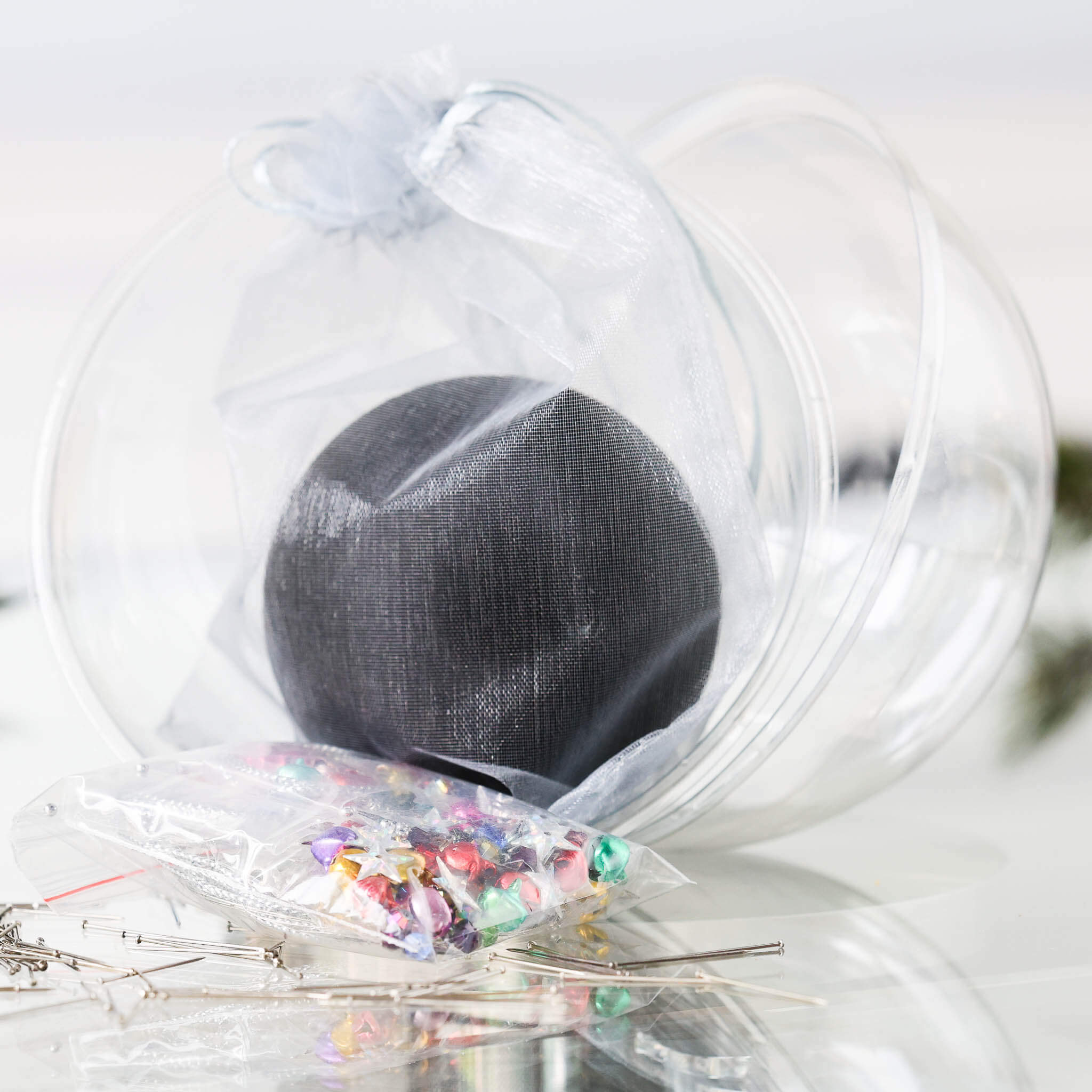 Push Pin Ornament Kit comes in a clear plastic container with  a black satin ball, tiny colorful jingle bells, and star sequins in a small clear bag, set against a white background.