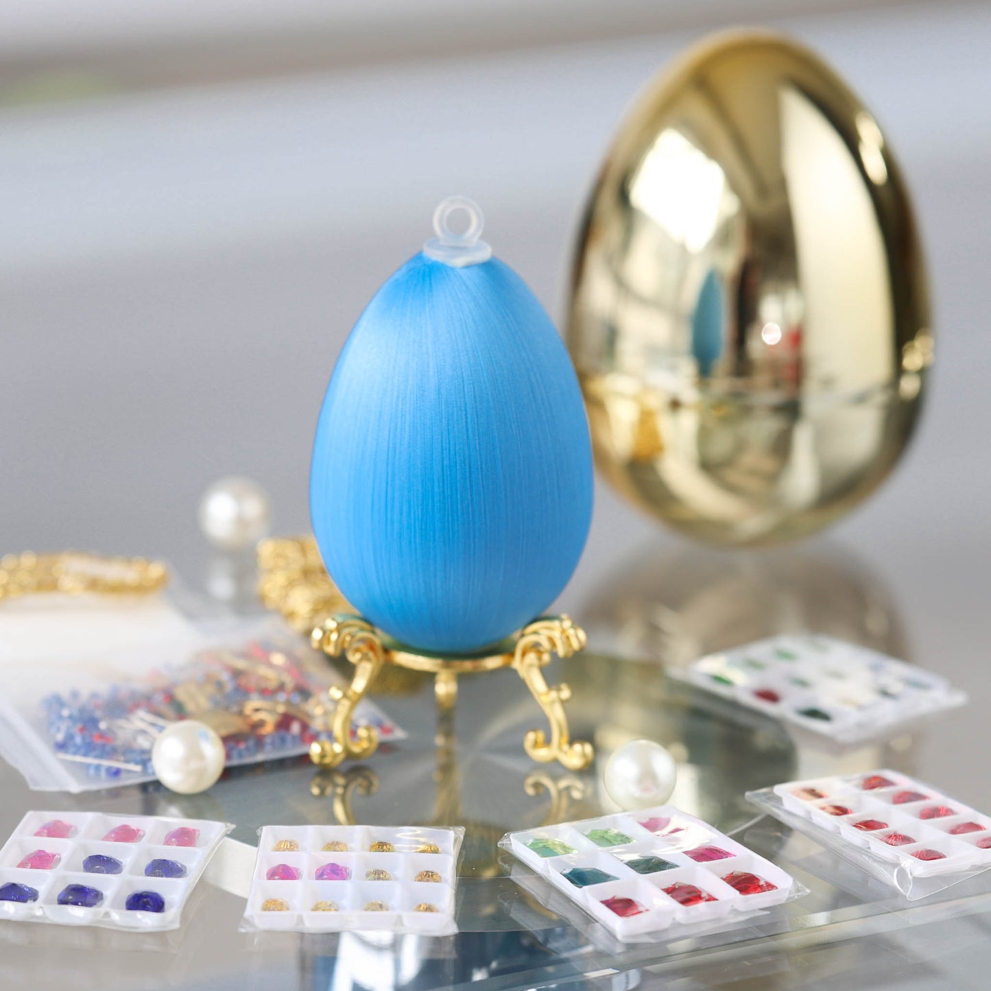 Sky blue satin foam egg on a stand with small packages, containing glass rhinestones for a push pin DIY Faberge inspired ornament project on a reflective surface by Whim5seasons.