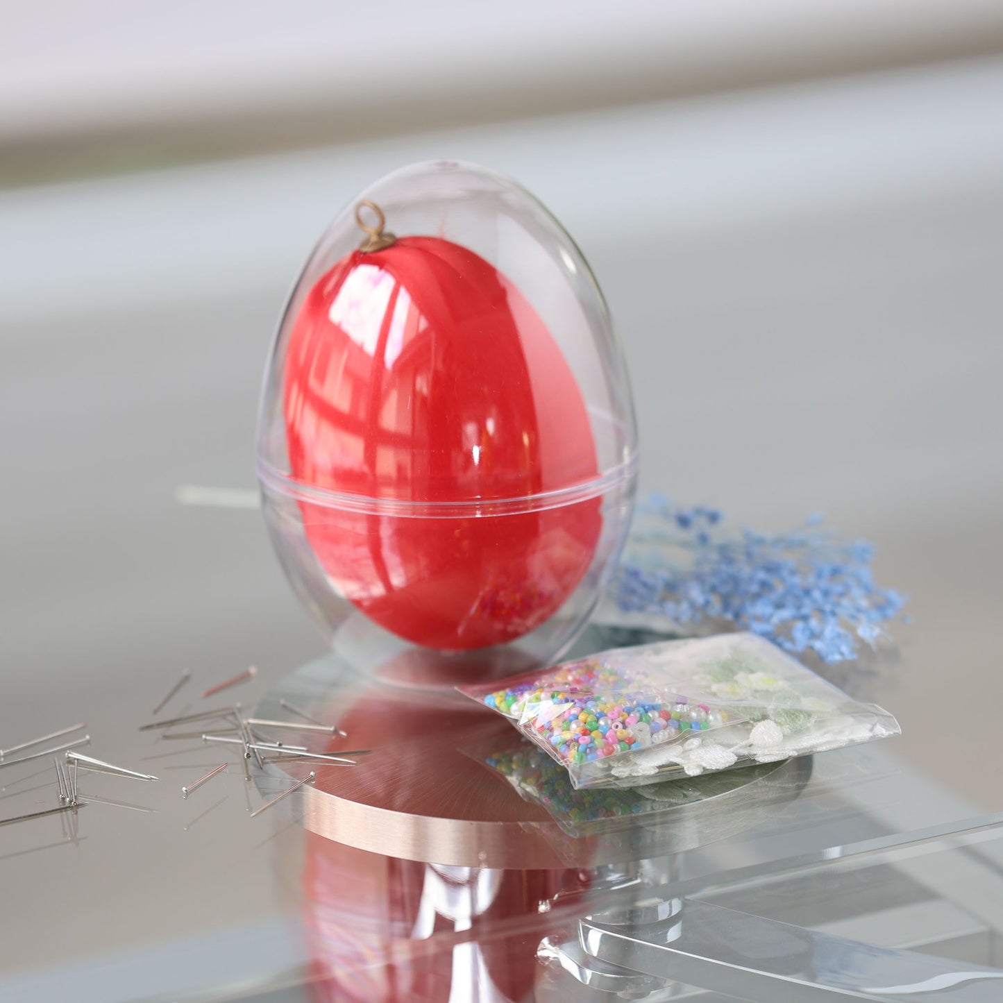 Clear plastic egg-shaped container with a large 4 inches red egg and a package of beads for a push pin project on a reflective surface by Whim5seasons.