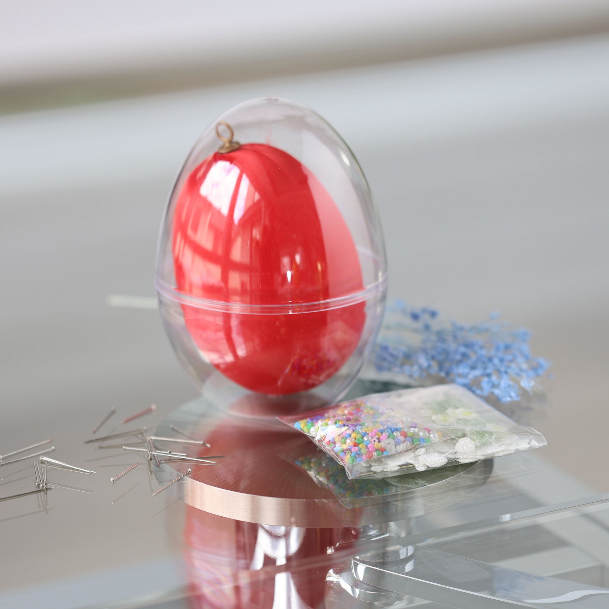 Clear plastic egg-shaped container with a large 4 inches red egg and a package of beads for a push pin project on a reflective surface by Whim5seasons.