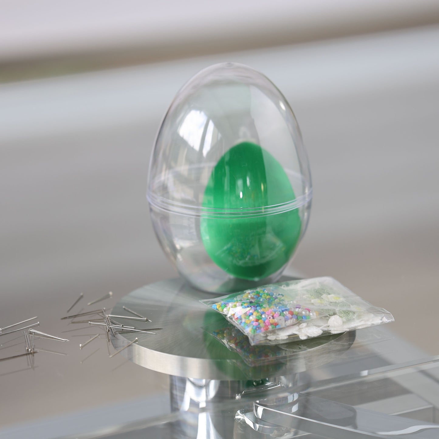 Clear plastic egg-shaped container with a satin green egg and a package of beads for a push pin project on a reflective surface by Whim5seasons.