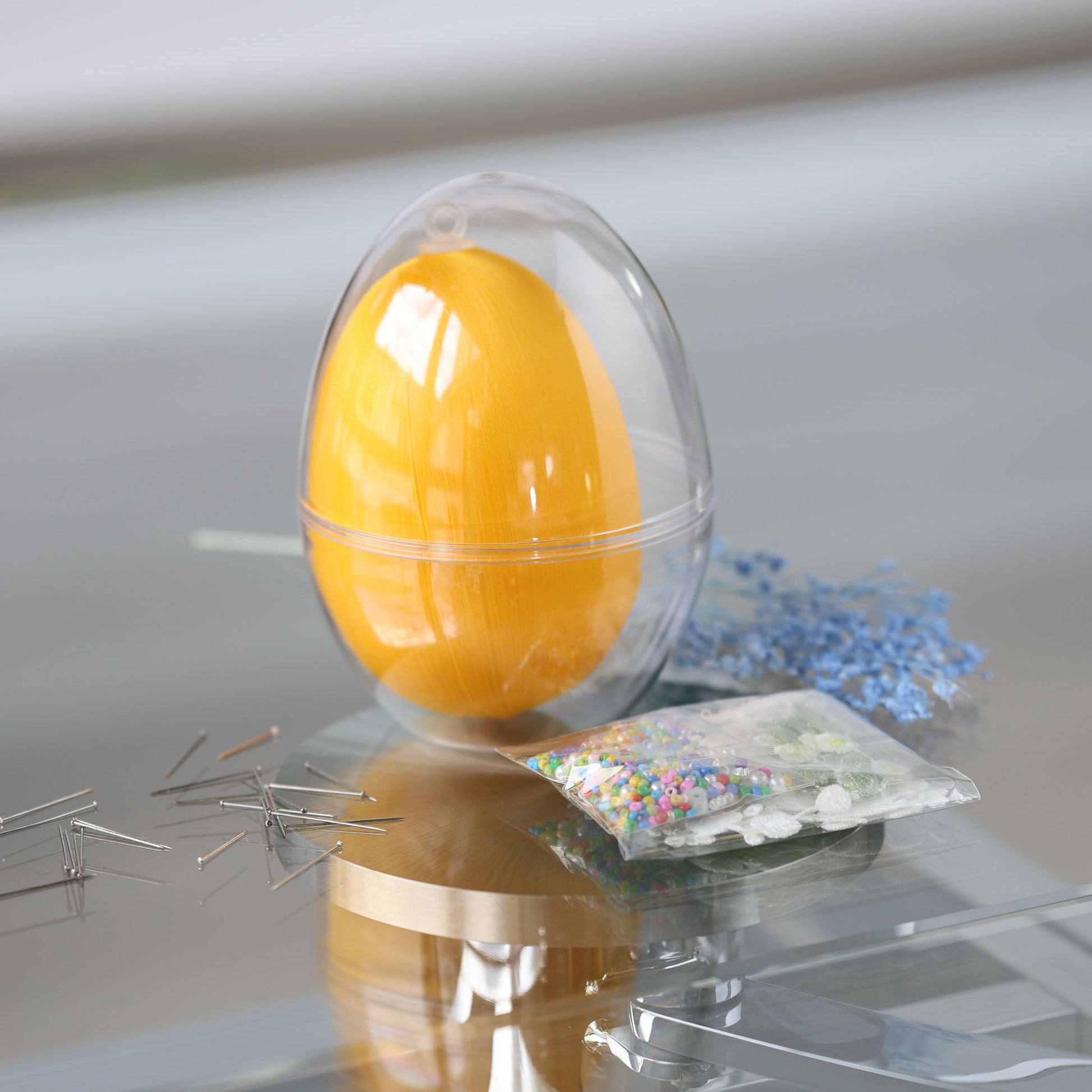 Clear plastic egg-shaped container with a satin bright yellow egg and a package of beads for a push pin project on a reflective surface by Whim5seasons.