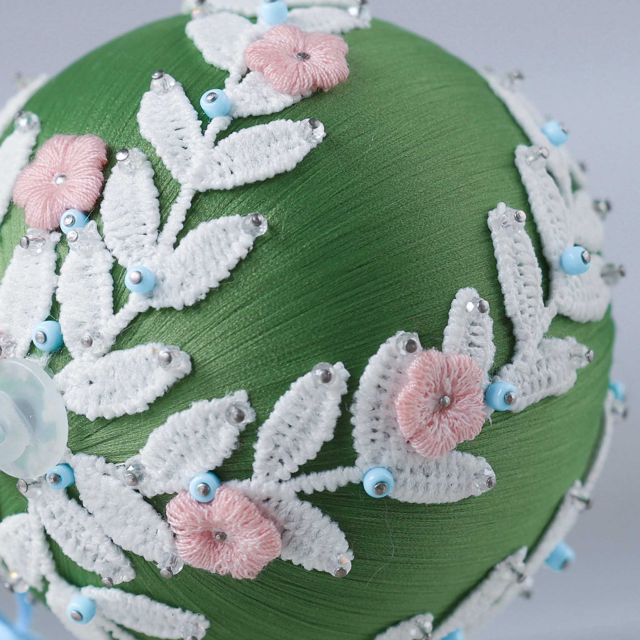 A close-up picture of a handmade Christmas Ornament Ball embellished with white floral trim, small blue beads, and pink embroidered flowers (Olive).