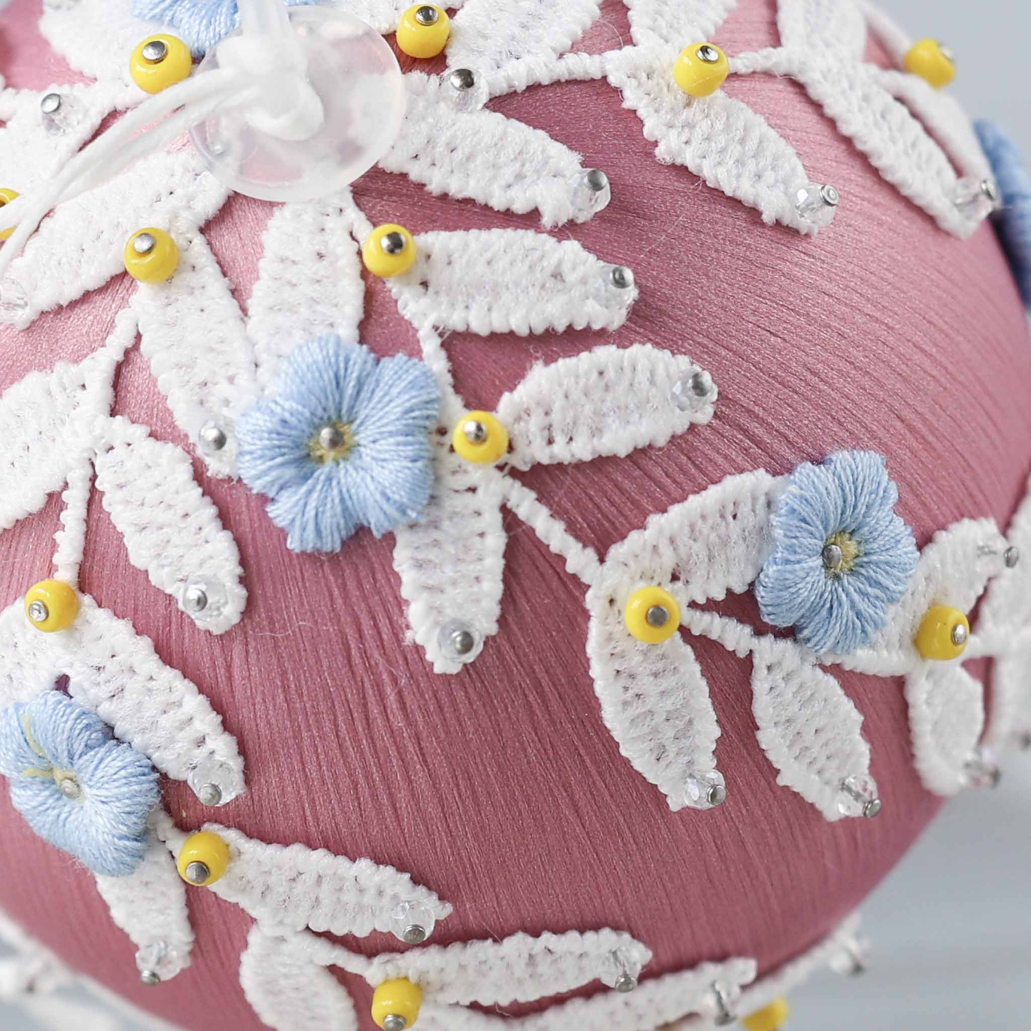 A close-up picture of a handmade Christmas or Easter beaded push pin Ornament embellished with small blue embroidered flowers, white trim, and yellow seed beads. (Pale Violet Red)