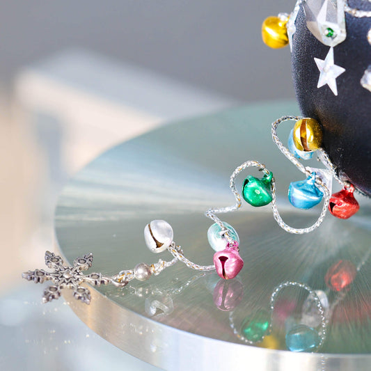 A close-up of a handmade push pin ornament's bottom with glass beads, tiny jingle bells, and metallic trim on a mirrored reflective platform.