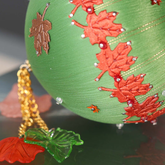 A close-up of a Handmade Push Pin Ornament in green featuring a rust colored maple ribbon trim pinned to a satin foam core with dressmaker pins, and some acrylic maple leaf charms. (Olive)
