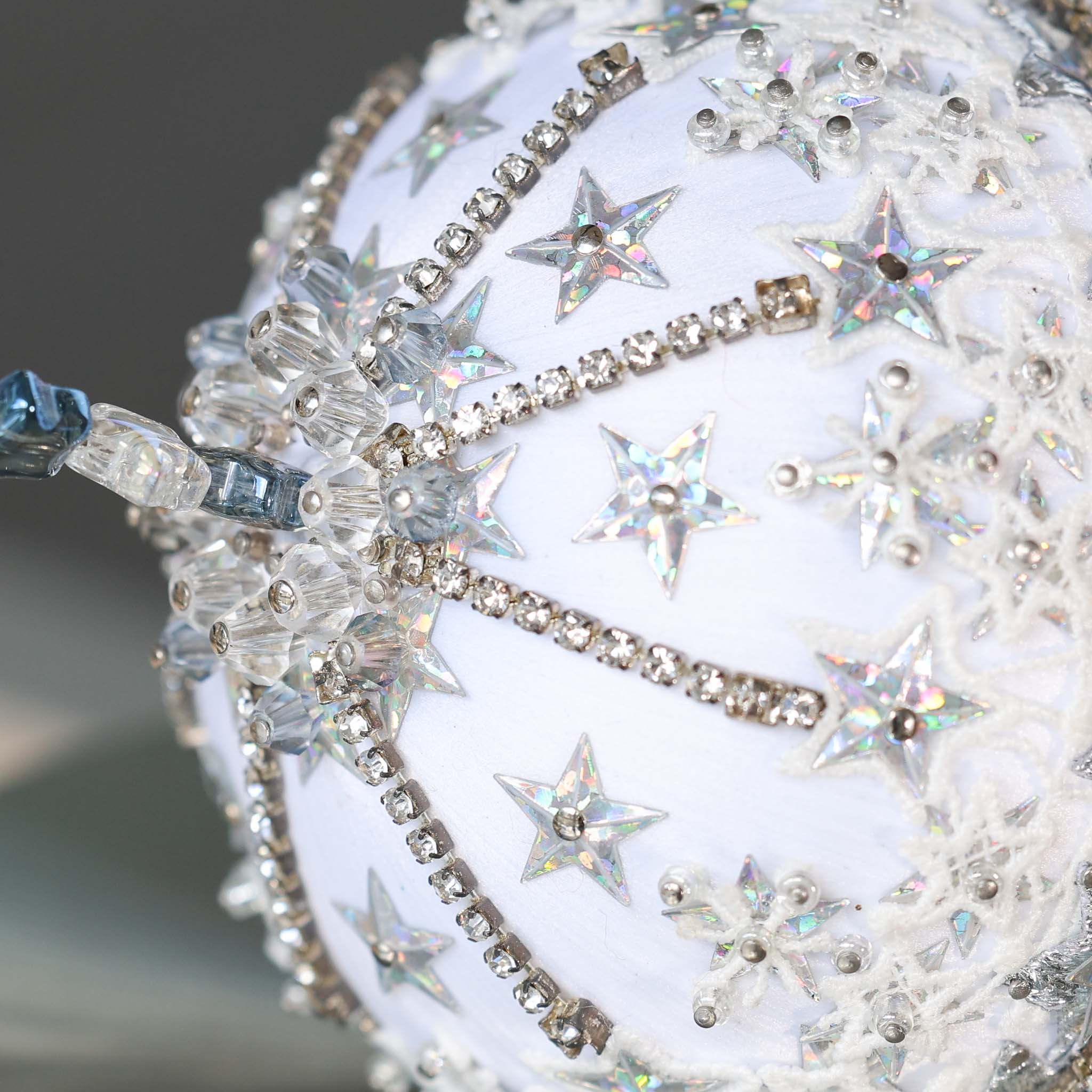 A close-up of sparkly beads and silver star-shaped sequins on a white satin foam core ball featuring a completed handmade push pin ornament. (White)