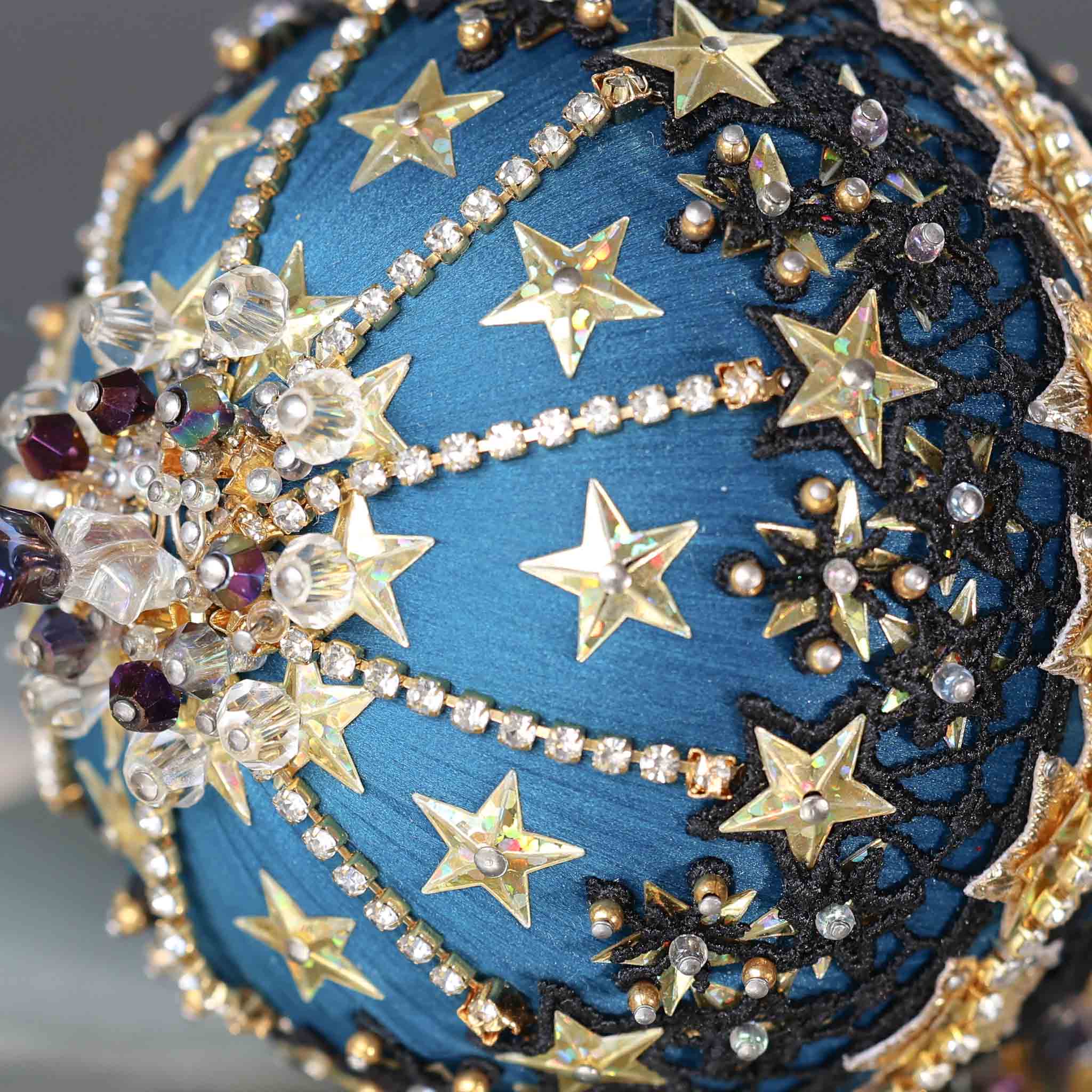 A close-up of dark purple beads and gold star-shaped sequins on a dark teal satin foam core ball featuring a completed handmade push pin ornament set against a dark gray background. (Dark Slate Blue)