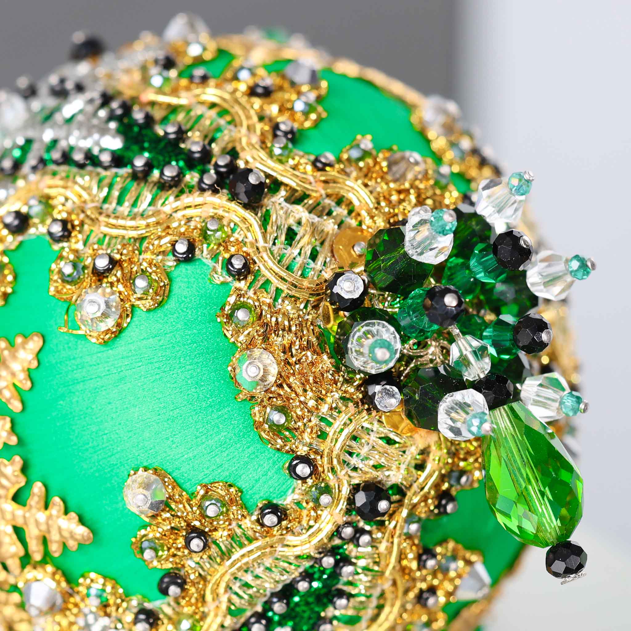 A close-up of a handmade push pin ornament's bottom with glass beads, and metallic trim on a satin ball with a gray background.   (Green)