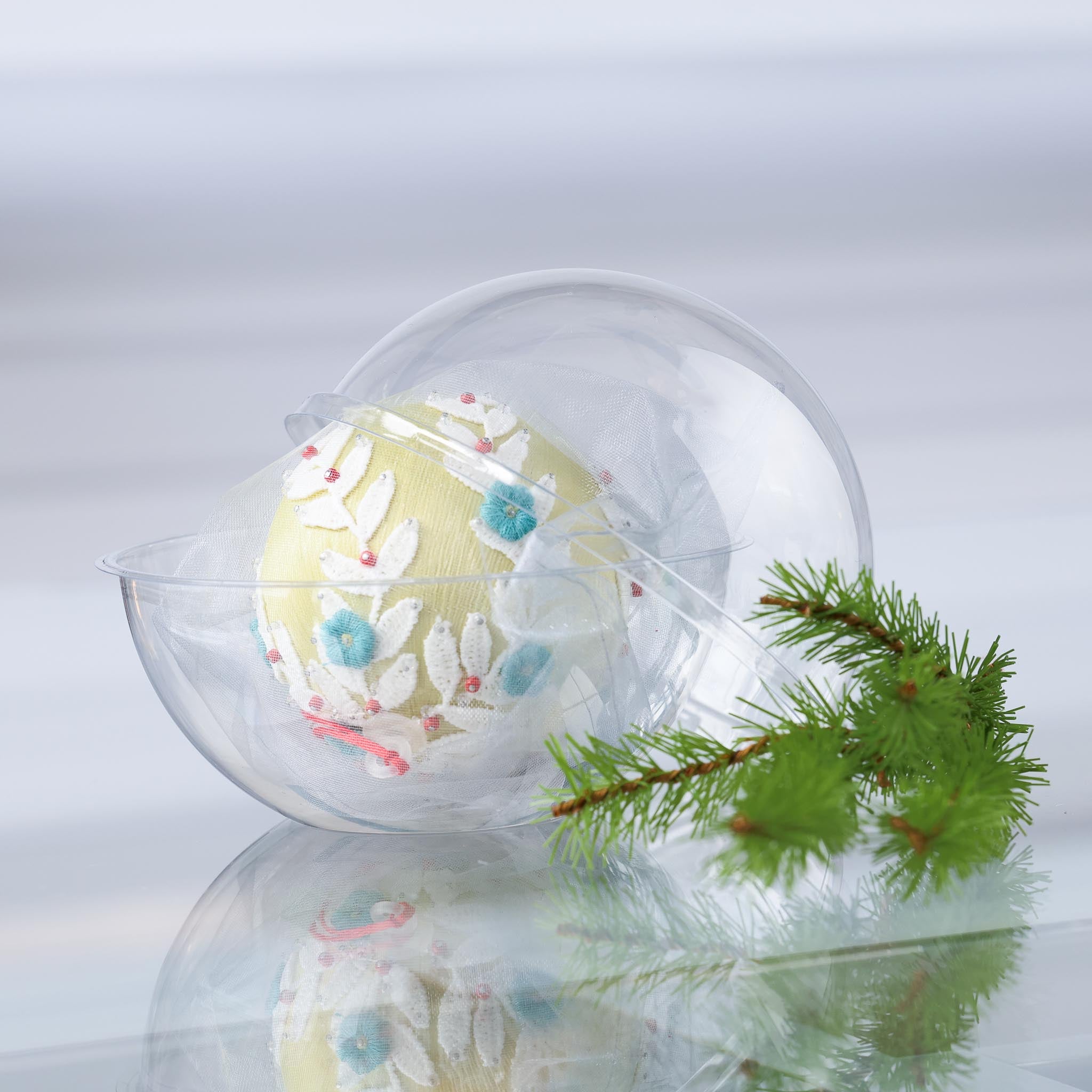 Satin push pin ornament Frosted Laurel in a clear storage container on a reflective surface with a holiday branch. (Khaki)