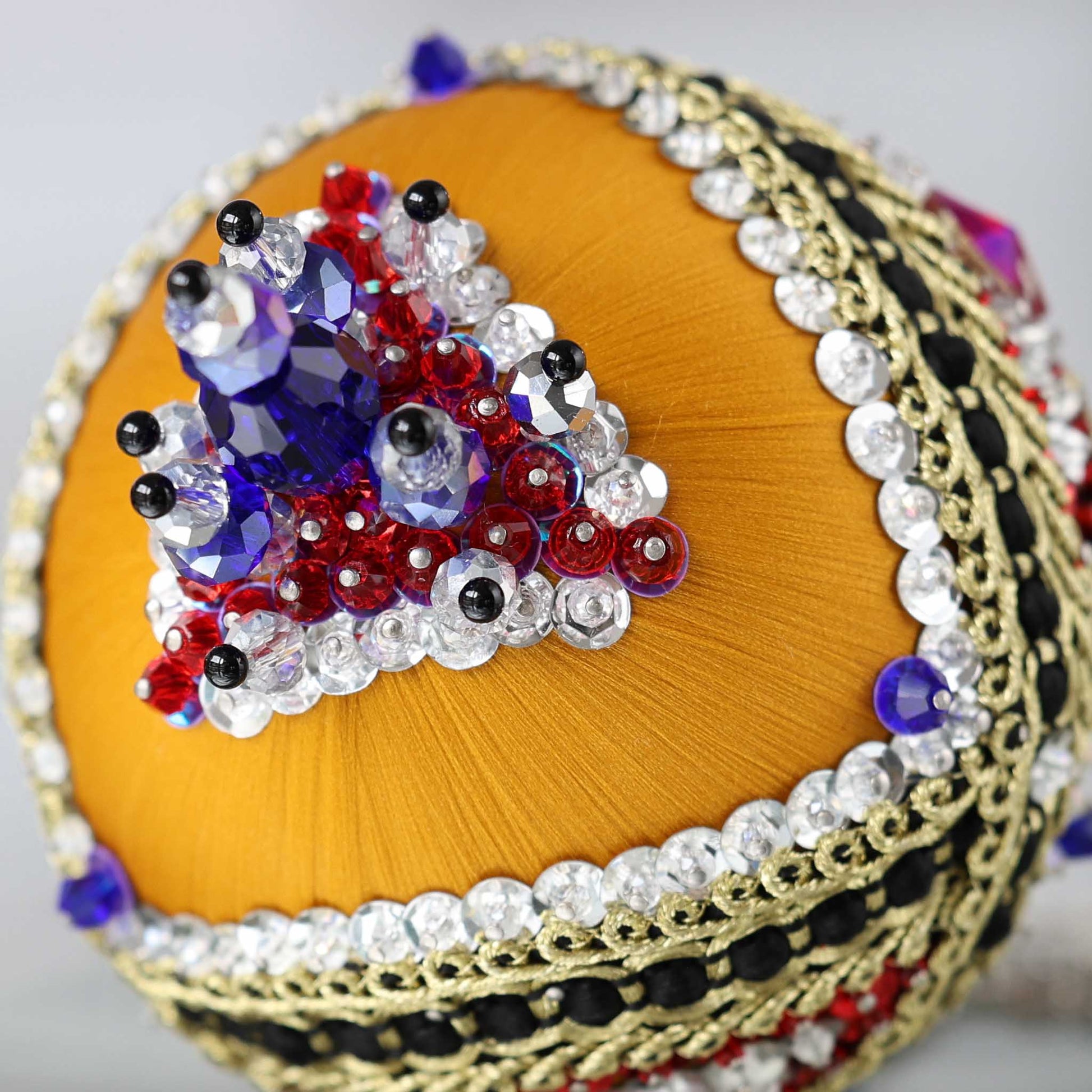 A close-up detail of a handmade satin push pin ornament featuring  red round rhinestones, gold trim, rows of silver sequins, and large blue beads attached with dressmaker pins by Whim5seasons.