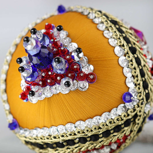 A close-up detail of a handmade satin push pin ornament featuring  red round rhinestones, gold trim, rows of silver sequins, and large blue beads attached with dressmaker pins by Whim5seasons.
