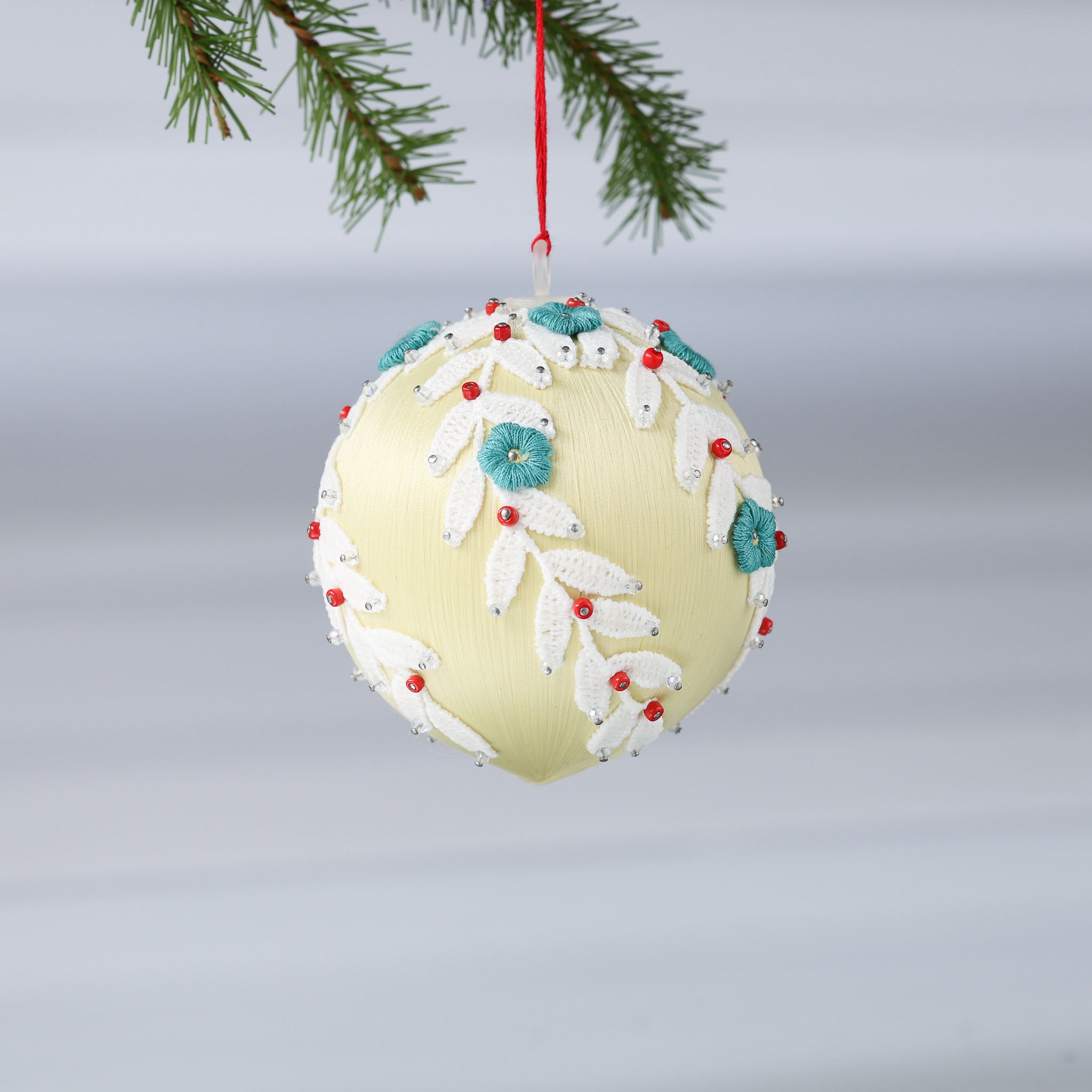 Handmade push pin beaded Christmas Ornament Ball embellished with embroidered flowers, white lace trim, and small clear beads hanging from a green evergreen branch. (Khaki)