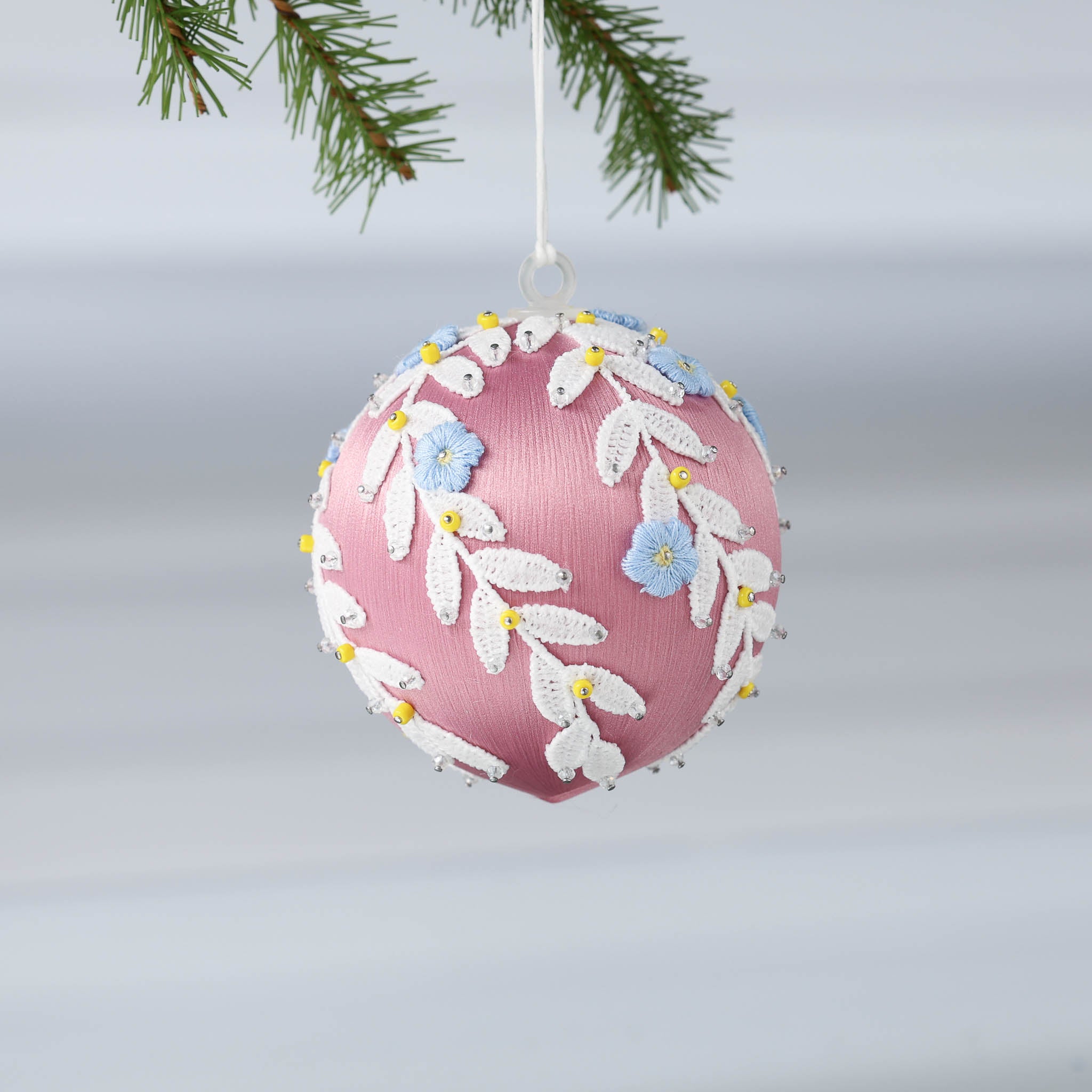 Handmade Christmas or Easter Beaded push pin Ornament Ball with blue embroidered flower patches, white trim, and beads pinned to a satin foam core and hanging on a light gray background.  (Pale Violet Red)