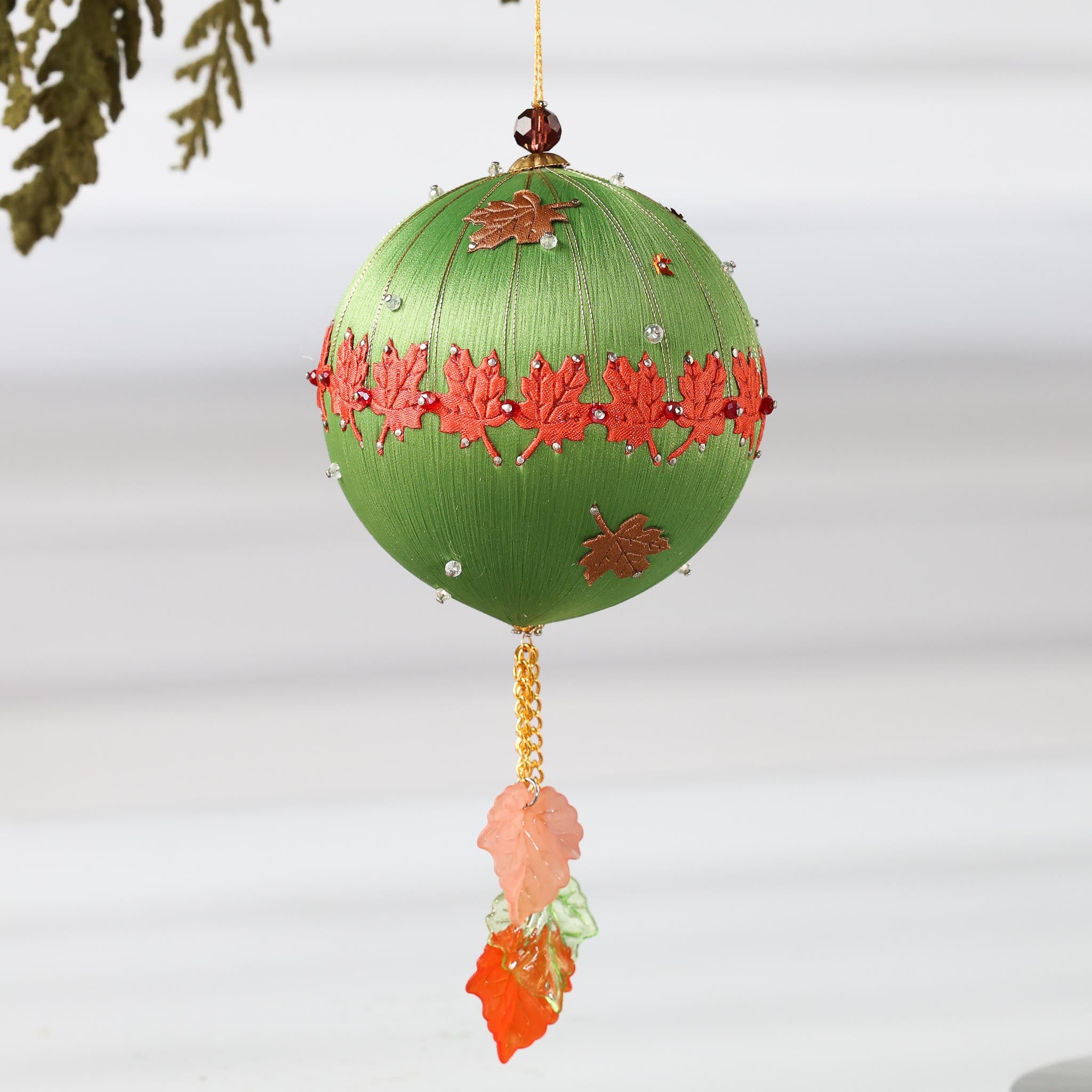 A Handmade Push Pin Ornament in olive featuring a rust colored maple ribbon trim pinned to a satin foam core with dressmaker pins, and some acrylic maple leaf charms, and clear seed beads. (Olive)