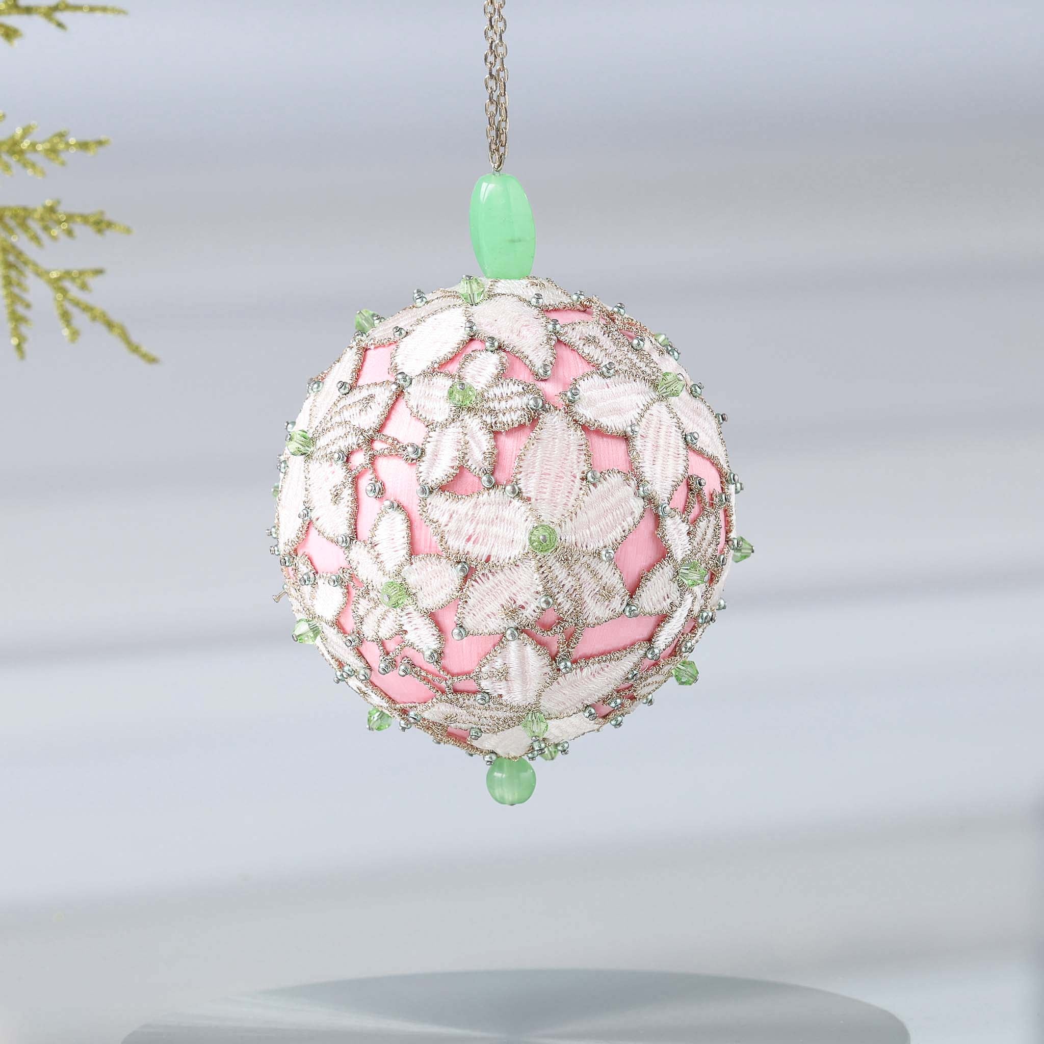 Handmade sequin & beaded pink satin ornament featuring flower trim hanging in front of gray background and holiday greenery by Whim5seasons.