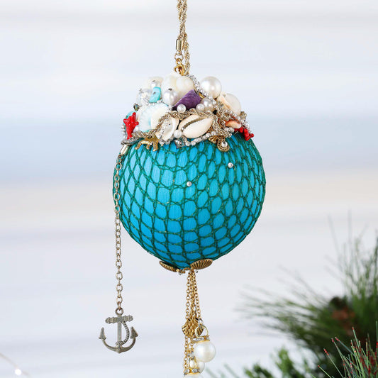 Handmade sequin & beaded teal-colored satin ornament featuring real shells and pearls hanging in front of gray background and holiday greenery by Whim5seasons.
