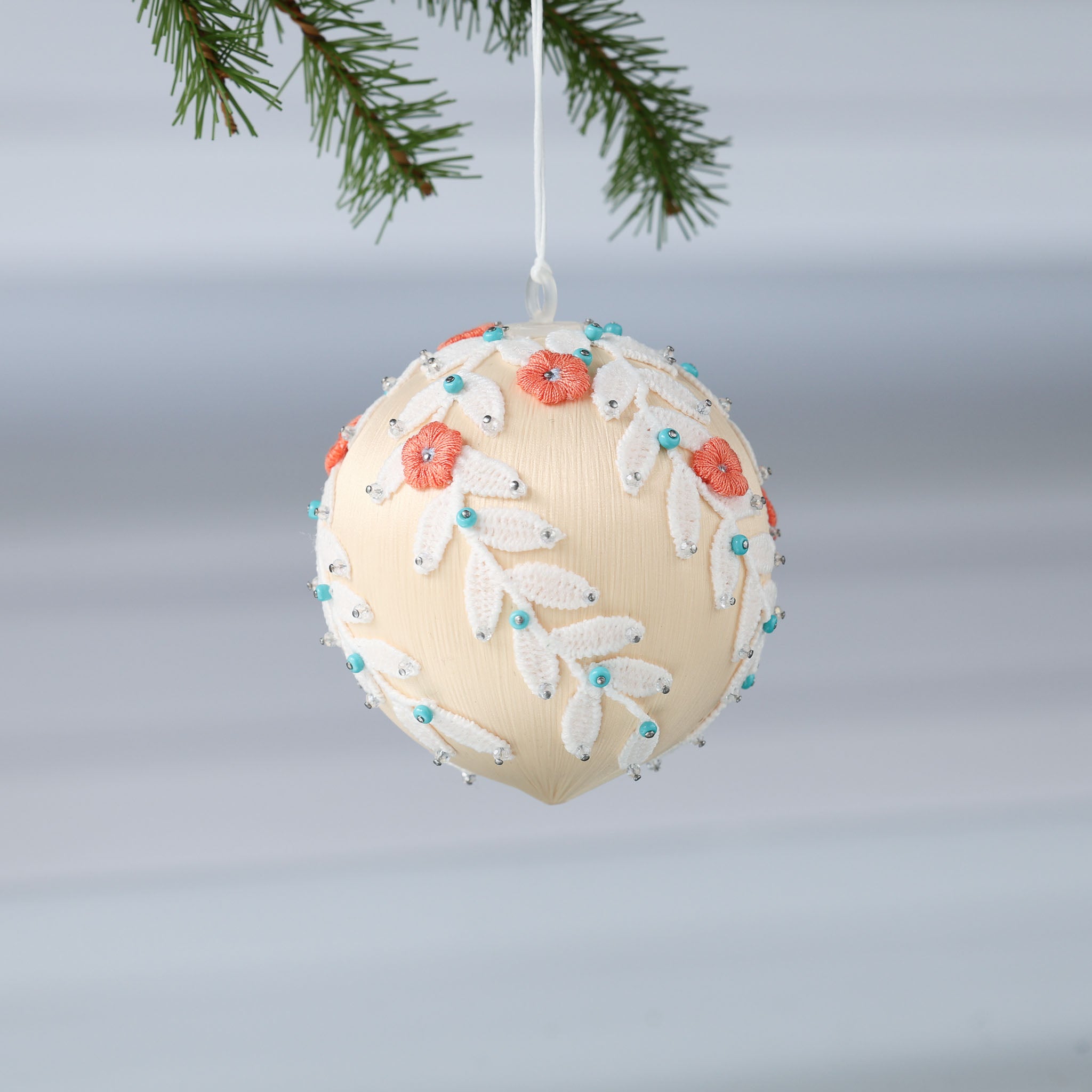 Finished Mid-Century Style Frosted Laurel Satin Ornament in a light tan color with orange embroidered flowers pinned to a satin foam core on a reflective surface. (Papaya Whip)