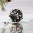 Load image into Gallery viewer, Decorative handmade black satin ornament with intricate designs on a white background on a reflective surface with a pinecone.
