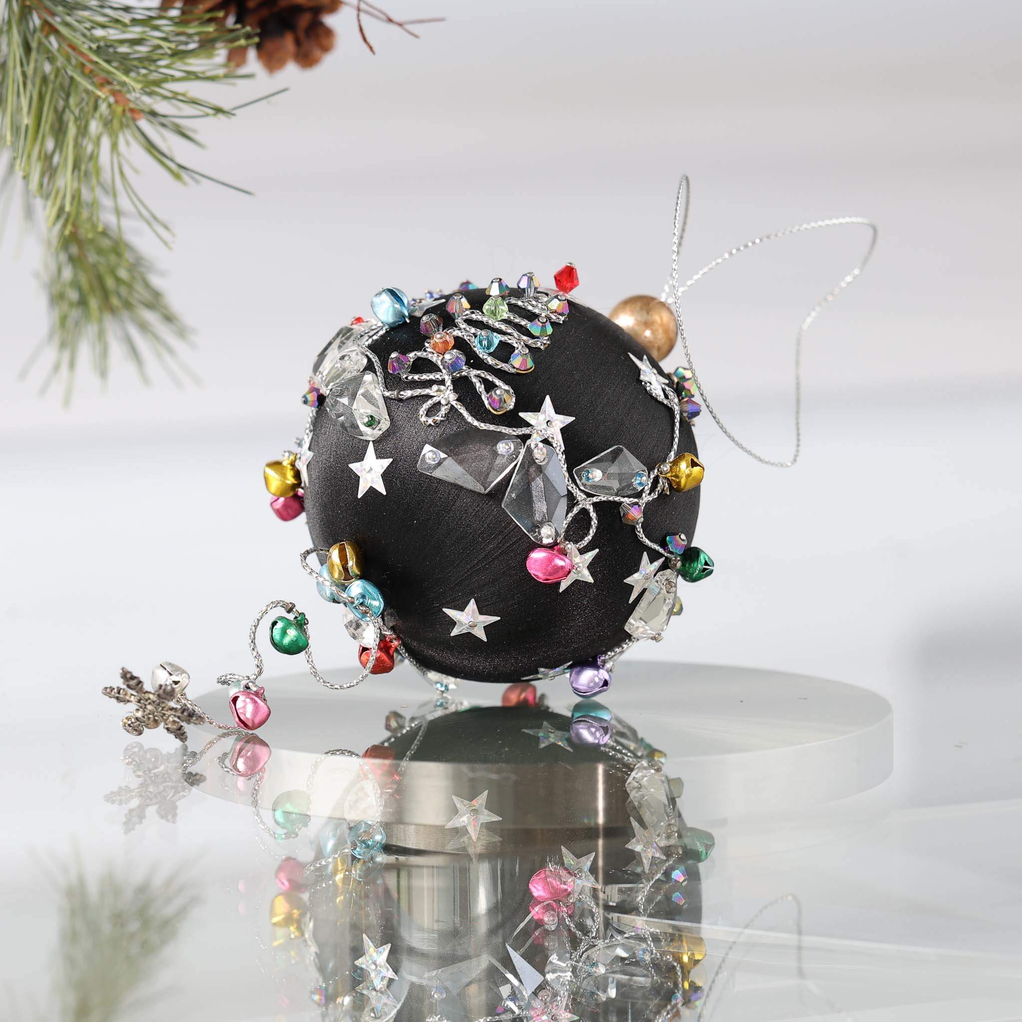 Decorative handmade black satin ornament with intricate designs on a white background on a reflective surface with a pinecone.