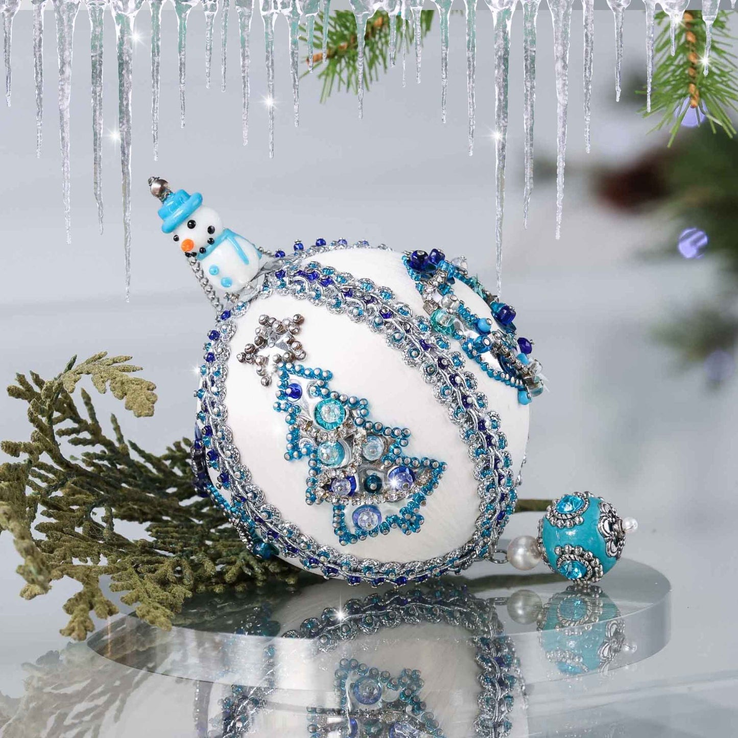 Handmade Christmas satin push pin ornament in white with blue and silver sequined tree, snowman bead, and a large Tibetan style bead on the bottom on a reflective surface by Whim5seasons.