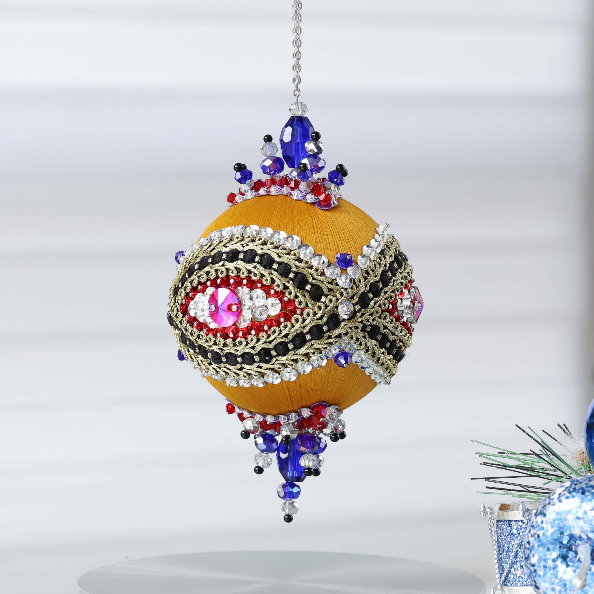 Handmade sequin & beaded satin ornament featuring  an eye design and red/blue beads combination hanging in front of gray background and holiday greenery by Whim5seasons.
