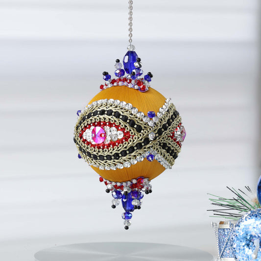 Handmade sequin & beaded satin ornament featuring  an eye design and red/blue beads combination hanging in front of gray background and holiday greenery by Whim5seasons.